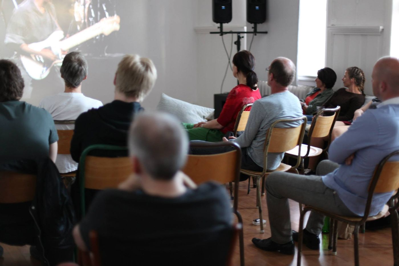 Publikum på stoler ser en konsertfilm projisert på veggen i et lyst lokale