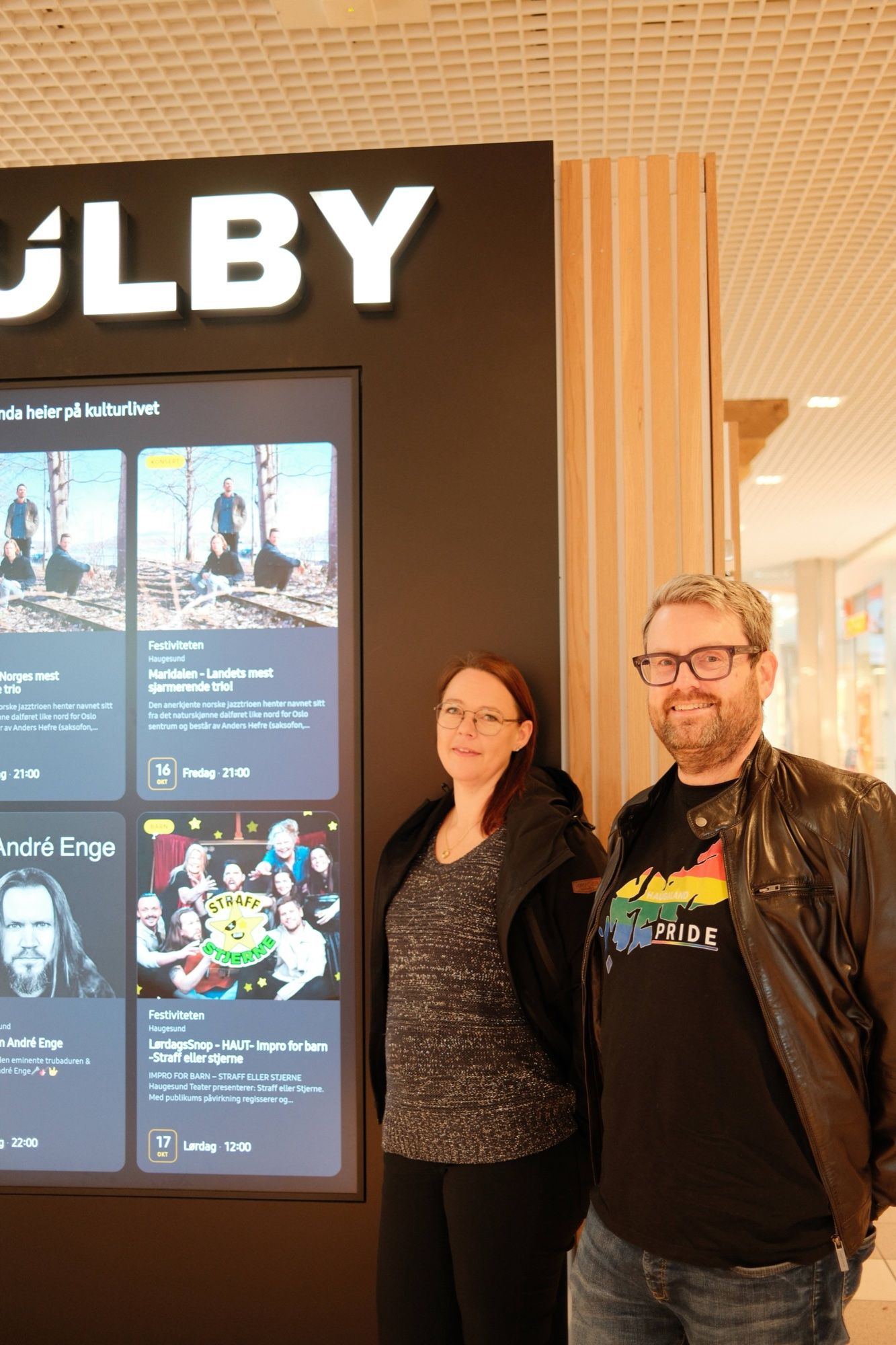 To personer, den ene i Haugaland Pride-t-skjorte, foran Kulby-skjermen med kulturprogrammet i Haugesund