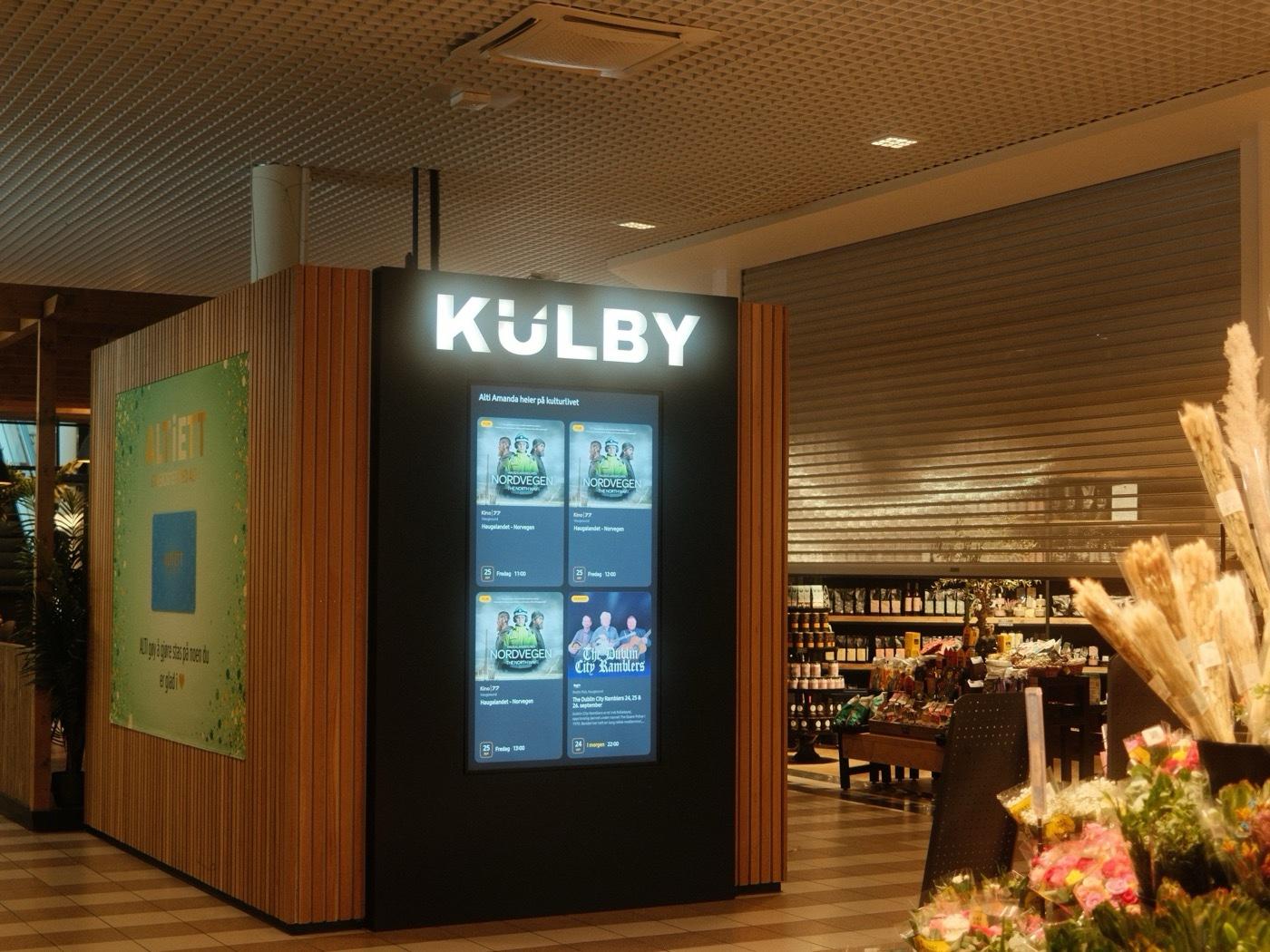 Kulby-kiosken i kjøpesenteret Alti Amanda i Haugesund: lyst KULBY-skilt over en skjerm med ukens program, blomster i forgrunnen